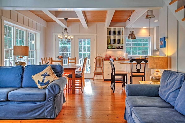 A spacious living area with a wooden floor, a blue sofa with an anchor pillow, a dining table with chairs, and a kitchen area with shelves and a hanging lamp.