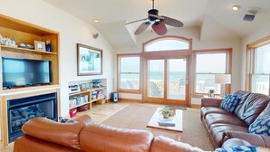 A spacious living room with a fireplace, a television, a bookshelf, a leather sofa, and a large window offering a view of the ocean. - Seaside | 6BR 4x4 OBX | Hot Tb | Sleeps 14 | 4 night stays available for fall (Carova)