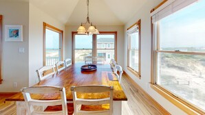 A dining room with a wooden table and chairs, large windows with beach views, and a hanging light fixture. - Seaside | 6BR 4x4 OBX | Hot Tb | Sleeps 14 | 4 night stays available for fall (Carova)