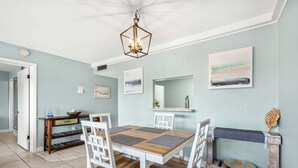 A dining area with a wooden table and white chairs, a hanging light fixture, and artwork on the walls. - Tropical Gem in Cocoa Beach W/ Private Balcony & Oceanfront Views! 🍹 (Cocoa Beach)