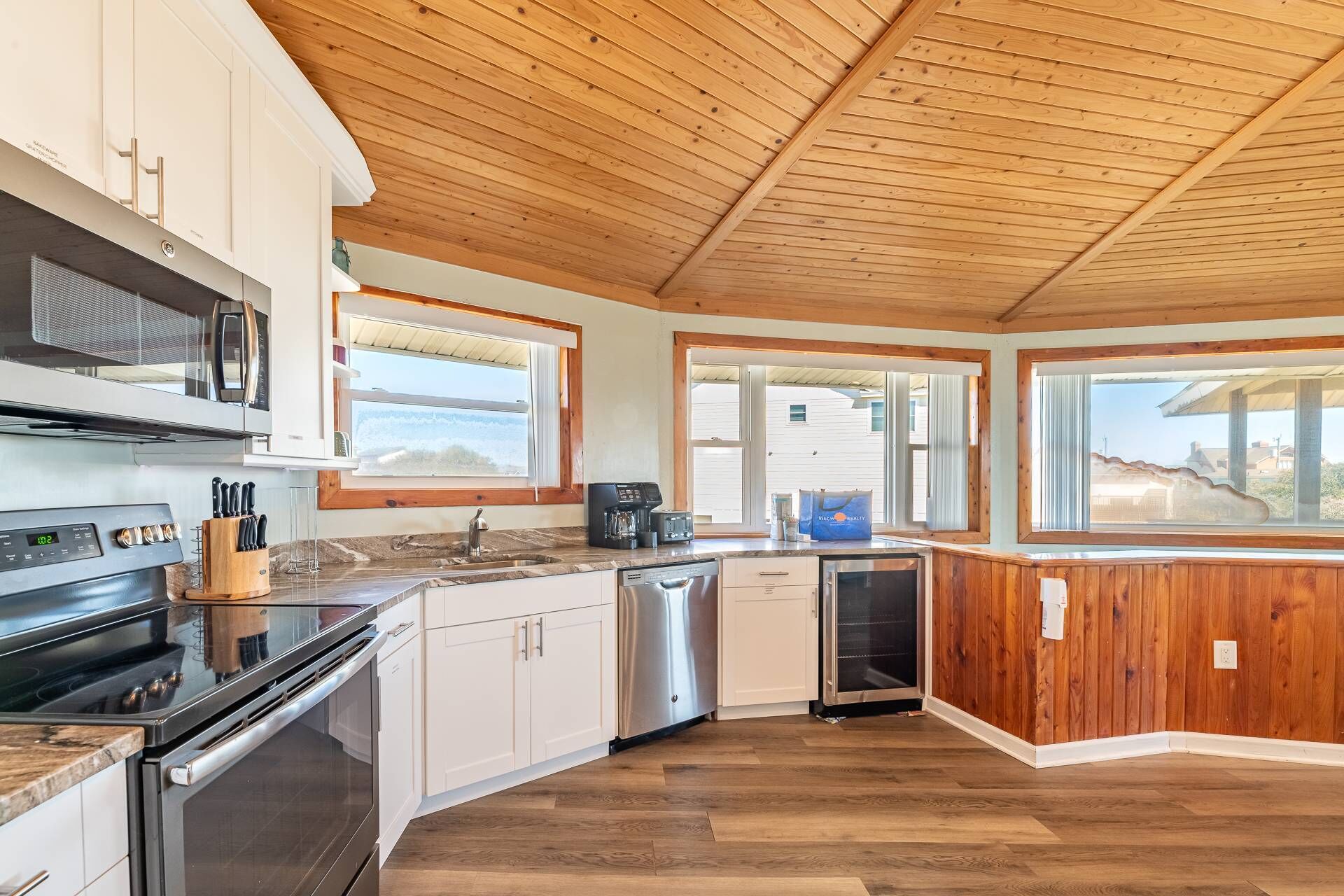A modern kitchen with wooden ceiling, white cabinets, stainless steel appliances, and large windows offering views.