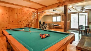 Una mesa de billar en una sala con suelo de madera, una escalera y un área de descanso al fondo.