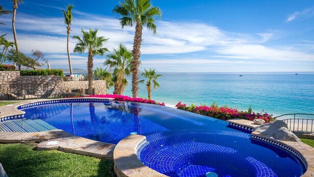 A infinity pool with a clear view of the ocean, surrounded by palm trees and a stone wall.