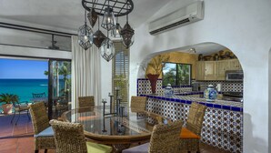 A dining area with a glass table and wicker chairs, a kitchen with blue and white tiles, and a view of the ocean.