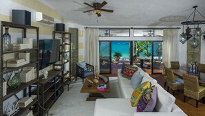 A spacious living area with a white sofa, a wooden coffee table, and a bookshelf. There is a ceiling fan, a view of the ocean through large windows, and outdoor furniture.