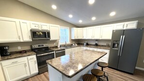 Uma cozinha moderna com armários brancos, eletrodomésticos de aço inoxidável, bancada de granito e uma ilha central com banquetas.