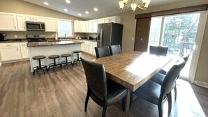 Uma cozinha moderna com armários brancos, uma ilha central com banquetas, uma mesa de jantar com cadeiras e um lustre.