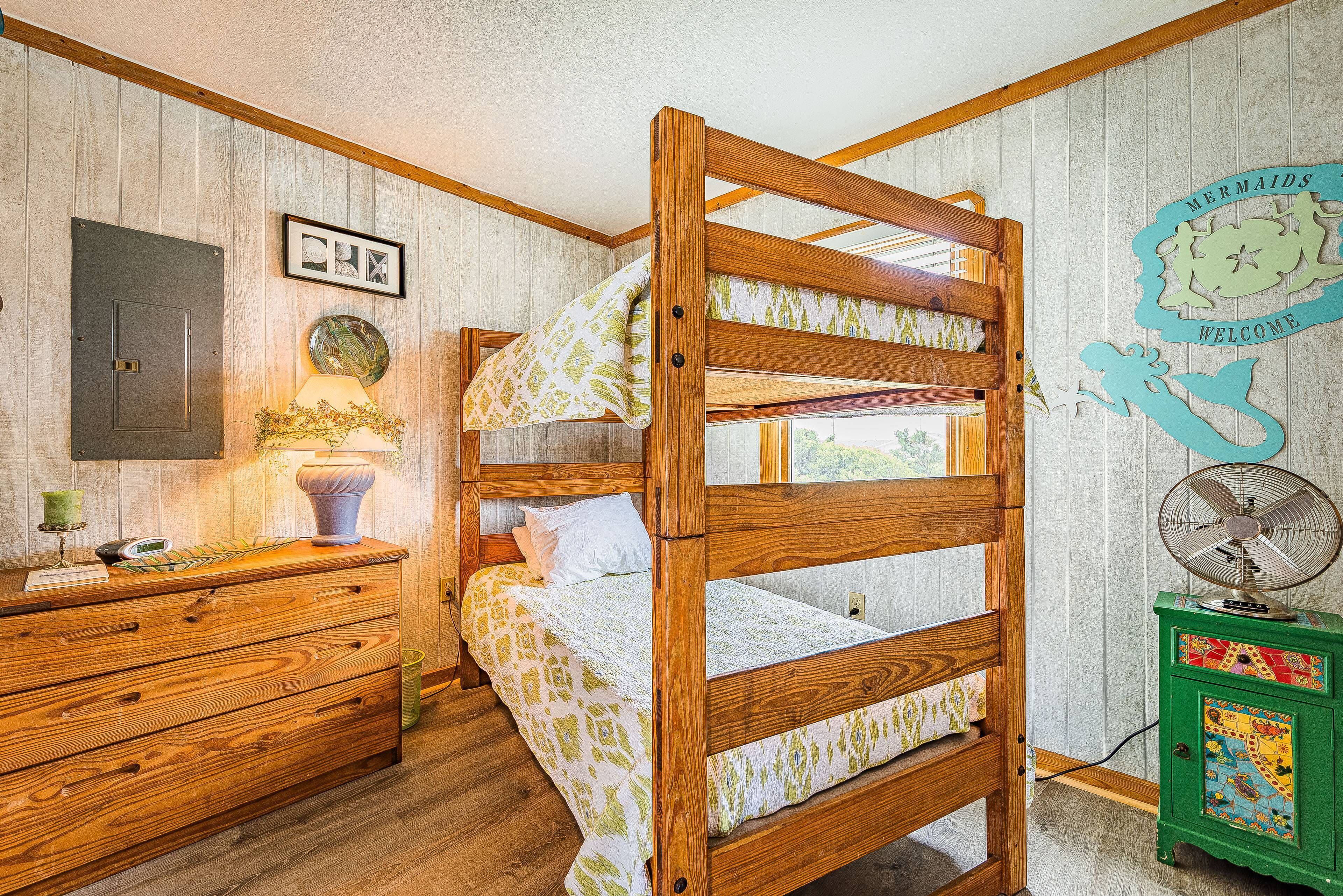 Una cama litera con estructura de madera y ropa de cama estampada, un aparador de madera con cajones, un mueble verde pequeño y un ventilador fijado a la pared.