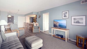 Una sala de estar con sofá, sillón y reposapiés, un televisor sobre un mueble bajo, y una zona de comedor con sillas y una mesa.