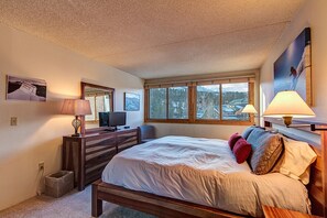 Un dormitorio con una cama grande, un aparador, un televisor y una ventana que ofrece vistas a árboles y edificios.