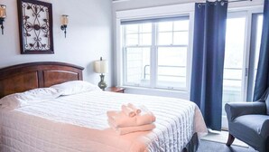 Quarto de hotel com uma cama grande, uma cadeira, janela com cortinas e arandelas na parede.