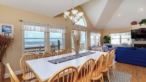 Un comedor con una mesa, sillas y una araña. Hay ventanas con cortinas, vista al océano y un televisor en la sala.
