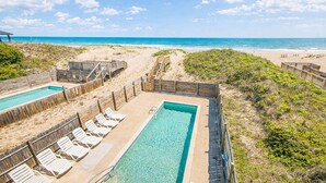 Zona de piscina con terraza de madera y sillas de descanso, rodeada de playa arenosa y vistas al océano.