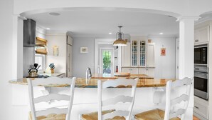 A kitchen with white cabinetry, a wooden island, and a dining area with white chairs and wicker cushions. - Walk to Beach and Town, Hot tub, Sleeps 8, Pet Friendly (Chatham)