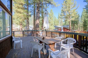 Une terrasse en bois avec une table et des chaises, entourée de grands arbres et sous un ciel dégagé.