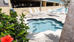 Un área de piscina con sillones de descanso y un jacuzzi, rodeado de follaje y un edificio con balcones.