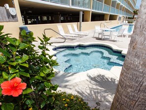 A pool area with lounge chairs and a hot tub, surrounded by greenery and a building with balconies.