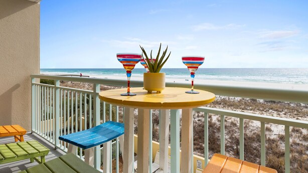 A balcony with a view of the beach, a small table with colorful glassware, and a potted plant.
