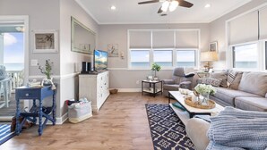A spacious living room with a ceiling fan, a television, and a view of the ocean through large windows. - Sweet Carolina - Perfect Surfside Oceanfront Location! (Surfside Beach)