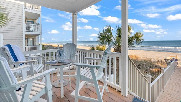 A covered deck with white railings, a table, and chairs, overlooking a beach with palm trees and the ocean. - Sweet Carolina - Perfect Surfside Oceanfront Location! (Surfside Beach)