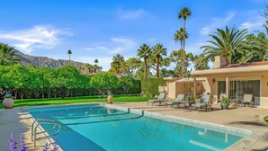 Una piscina con zona de patio, sillas de descanso y vistas a palmeras y montañas.