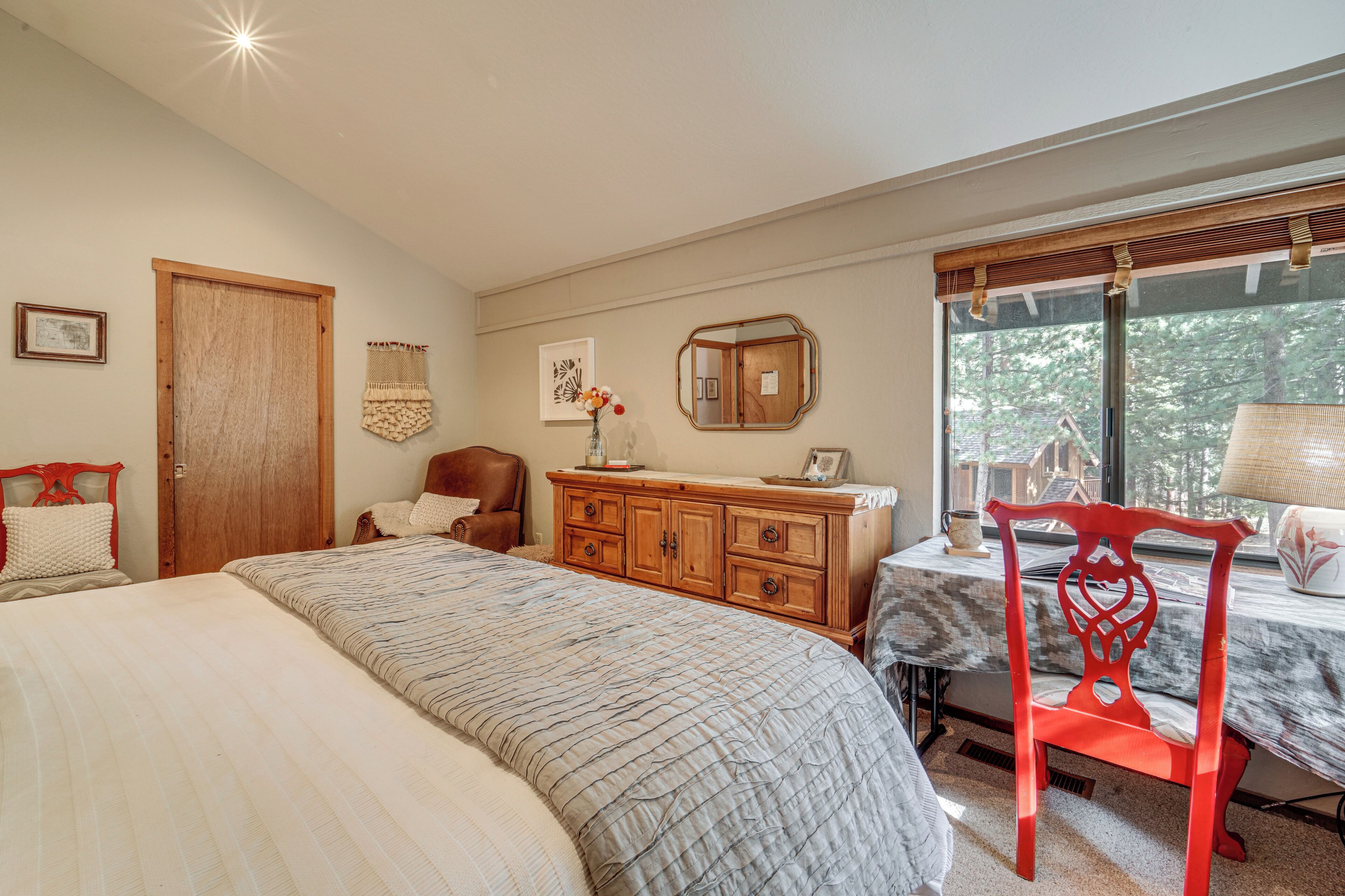 Un dormitorio con cama, cómoda de madera, una silla, una mesa y ventana con vista a árboles.