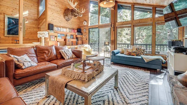 L’intérieur chaleureux d’un chalet, avec un grand plafond en bois, une tête de wapiti fixée au mur et une vue sur la forêt à travers de grandes fenêtres.