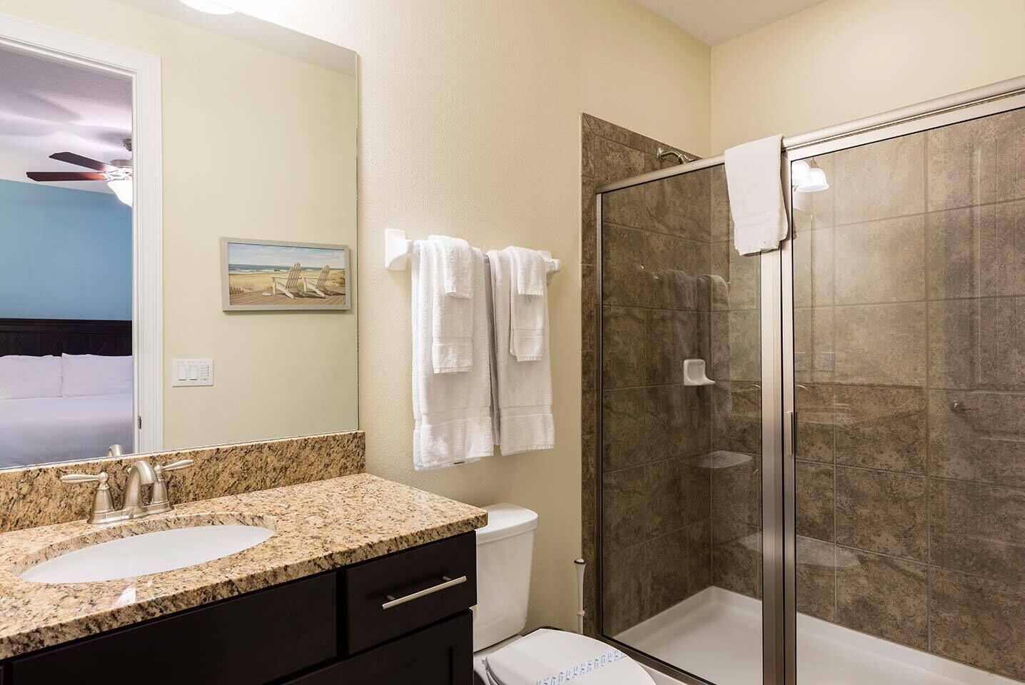 A bathroom with a granite countertop, a toilet, a shower with a glass door, and a bed visible through an open door.