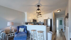 A modern kitchen with white cabinetry, a central island, and stainless steel appliances. There is a blue armchair and two white bar stools. - 910-B Turtle Cove: Beachfront Fun & Pool Access! (Surf City)