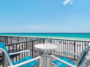 Une vue sur la plage depuis un balcon, avec une table blanche et des chaises bleues, donnant sur une plage de sable où l’on aperçoit des parasols et des gens.