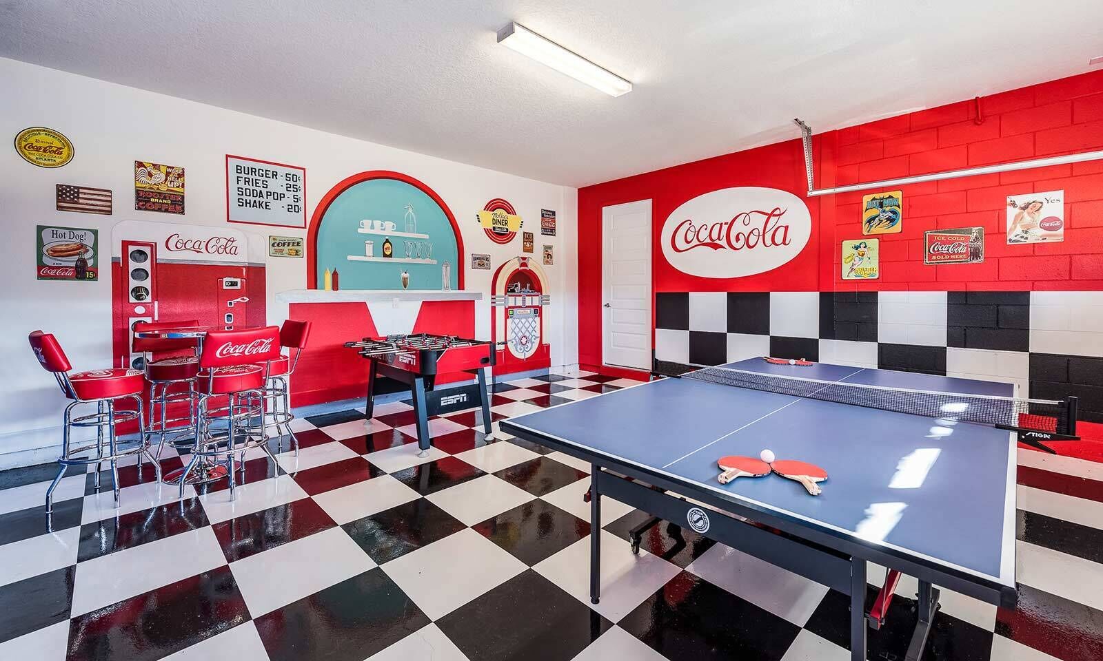 A retro-themed room with a foosball table, a ping pong table, and red Coca-Cola branded chairs.