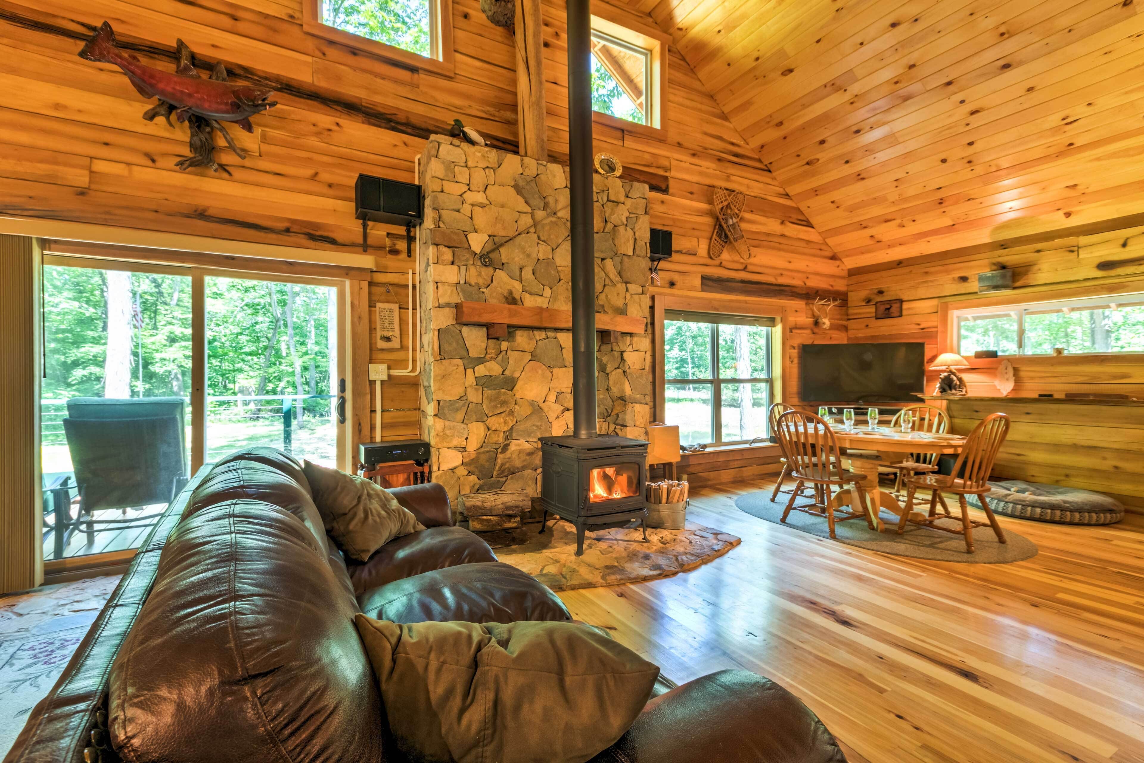 A cozy cabin interior with a stone fireplace, leather sofa, dining table, and large windows.