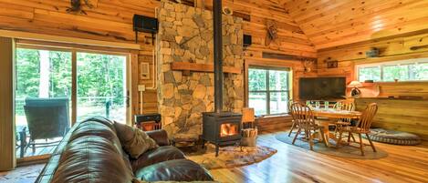 A cozy cabin interior with a stone fireplace, leather sofa, dining table, and large windows.