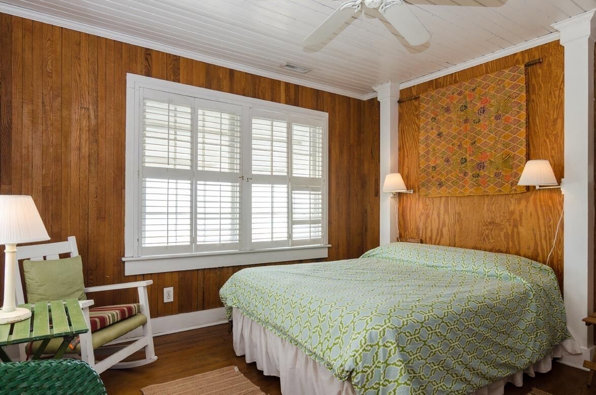 Een slaapkamer met houten wanden, een bed met een gestreept beddegoed, een schommelstoel en een raam met luiken.