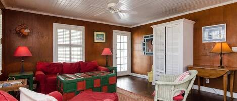 A cozy room with a red sofa, a green and red quilted coffee table, a wicker chair, and a desk with a lamp.