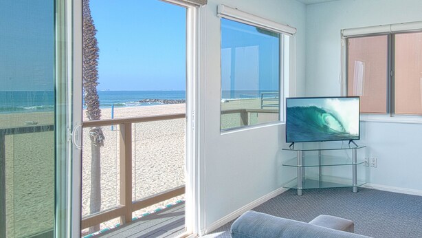 A room with large windows overlooking a beach, a television on a glass stand, and a blue couch. - A magnificent view awaits you as you enter...this upper unit is one-of-a-kind! (Newport Beach)