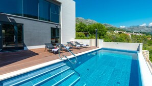 Un espace piscine moderne avec une terrasse en bois, des chaises longues et une vue sur les montagnes.