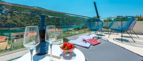 A table with a bottle of wine, glasses, a bowl of strawberries, and a menu on a balcony overlooking a scenic river and hills.
