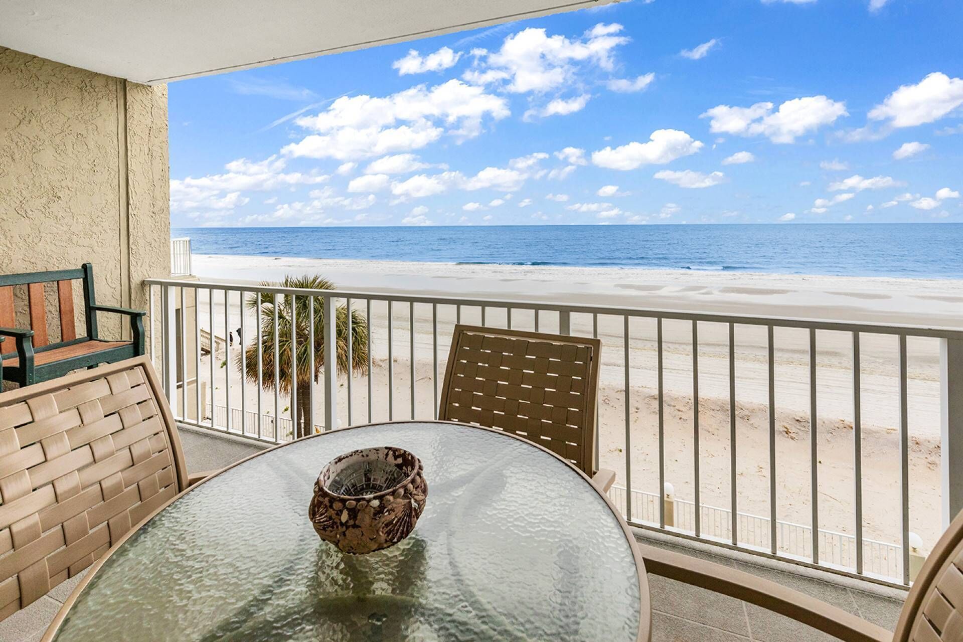 A balcony with a glass table, a chair, and a decorative bowl. The balcony overlooks a beach with a clear sky and ocean.