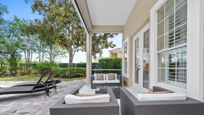 Uma área de estar coberta ao ar livre, com móveis de vime e uma mesa de vidro, com vista para um jardim ajardinado e um edifício residencial.