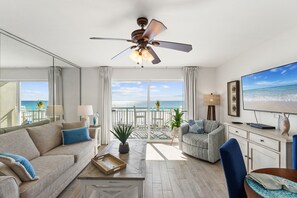 Een ruime woonkamer met een grote televisie, comfortabele zitplaatsen, een plafondventilator en een balkon met uitzicht op zee.
