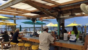 Een bar aan het water met houten stoelen en parasols, met uitzicht op een wateroppervlak.