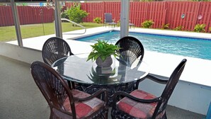 Een buitenpatio met een glazen tafel, omringd door rieten stoelen, een bloempot en een zwembad op de achtergrond.