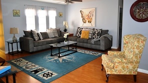 A living room with two grey sofas, a coffee table, a floral armchair, a blue rug with nautical symbols, and a ceiling fan.