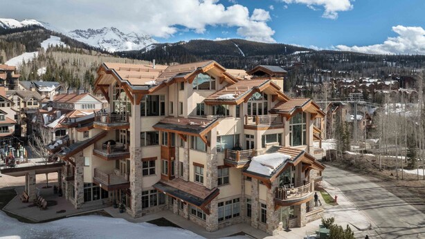 A multi-story hotel with balconies and large windows, set against a mountainous backdrop.