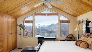 A bedroom with a large bed, wooden ceiling, and a view of mountains through large windows.