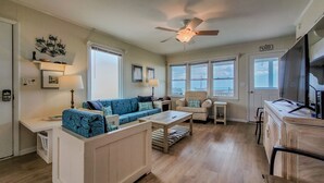 Uma sala de estar espaçosa com sofá, mesa de centro e um rack para TV. Há janelas que oferecem luz natural e vista para o exterior.