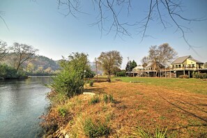 A riverside property with a grassy lawn, a wooden deck, and a building with a large window overlooking the water. - Peaceful Reliance Cabin w/ Deck on Hiwassee River! (Reliance)