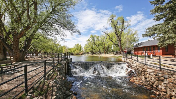 Una pequeña cascada en un parque con un muro de piedra, árboles y un edificio rojo al fondo.