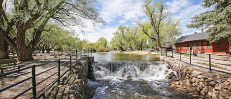 Una pequeña cascada en un parque con un muro de piedra, árboles y un edificio rojo al fondo.